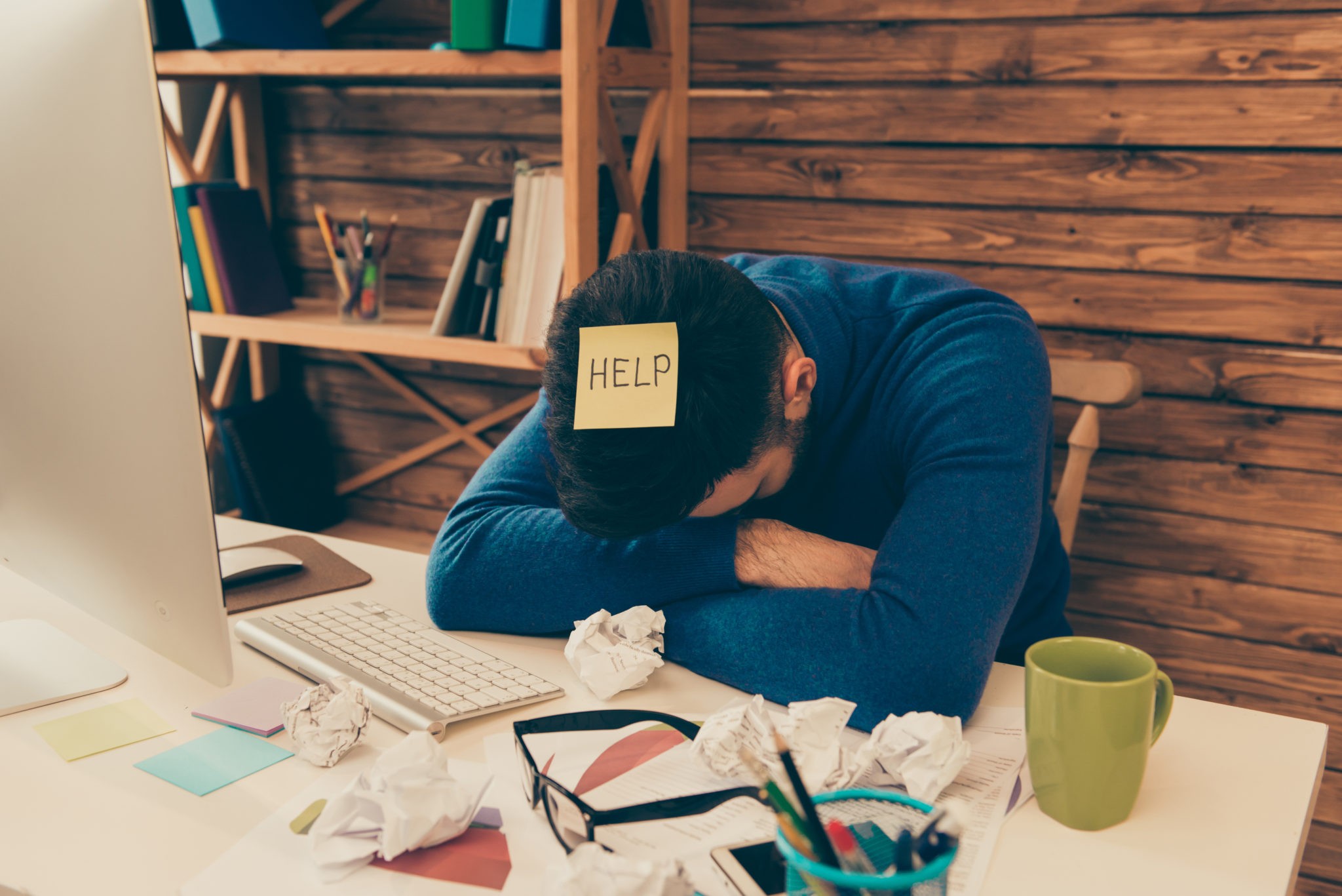 A man with his head down on his desk with a "help" sticky note