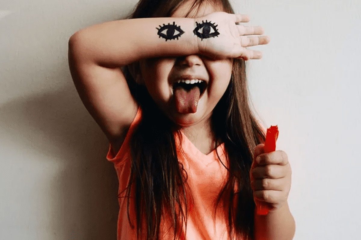 A young girl covers her eyes with her arm and sticks out her tongue. Two eyes are drawn on her arm over her own eyes.