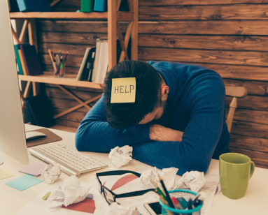 A man with his head down on his desk with a "help" sticky note